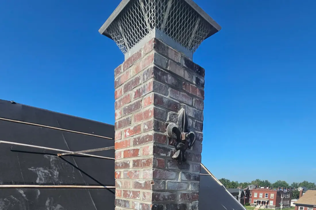 A rebuilt chimney topped with a cast cap and a mesh rain cap, a fleur de lis set into the brick face.