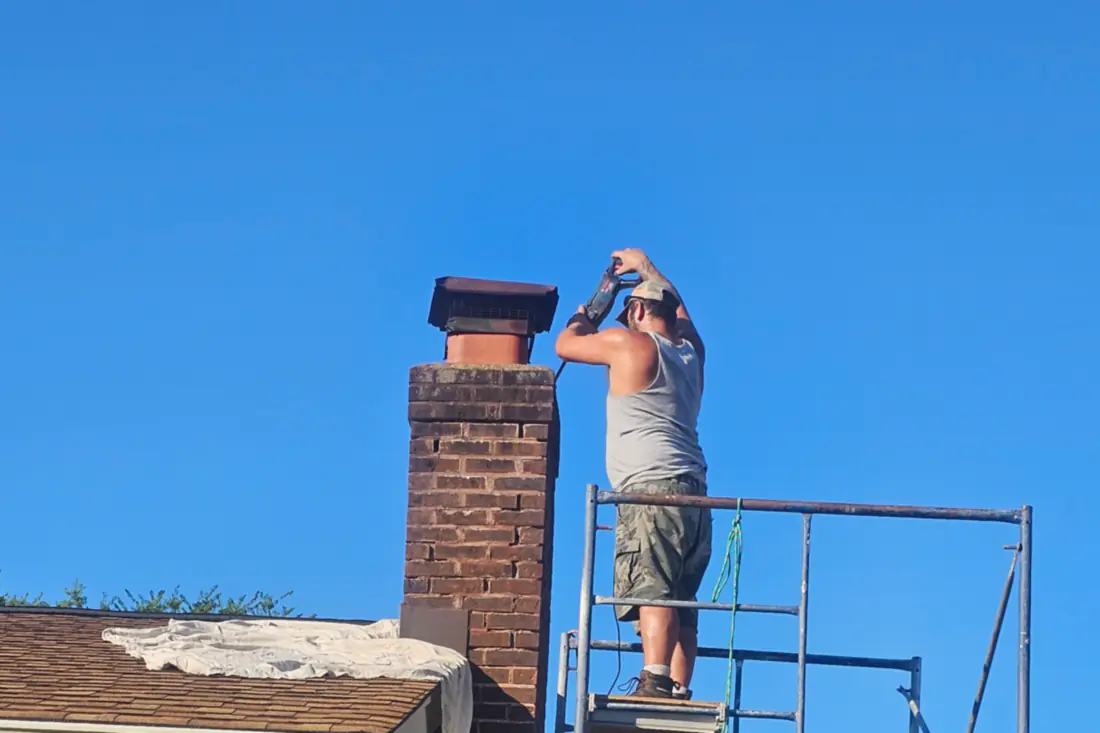On the scaffold, mid-rebuild: working the top of a brick chimney from a plank platform against a clear sky.